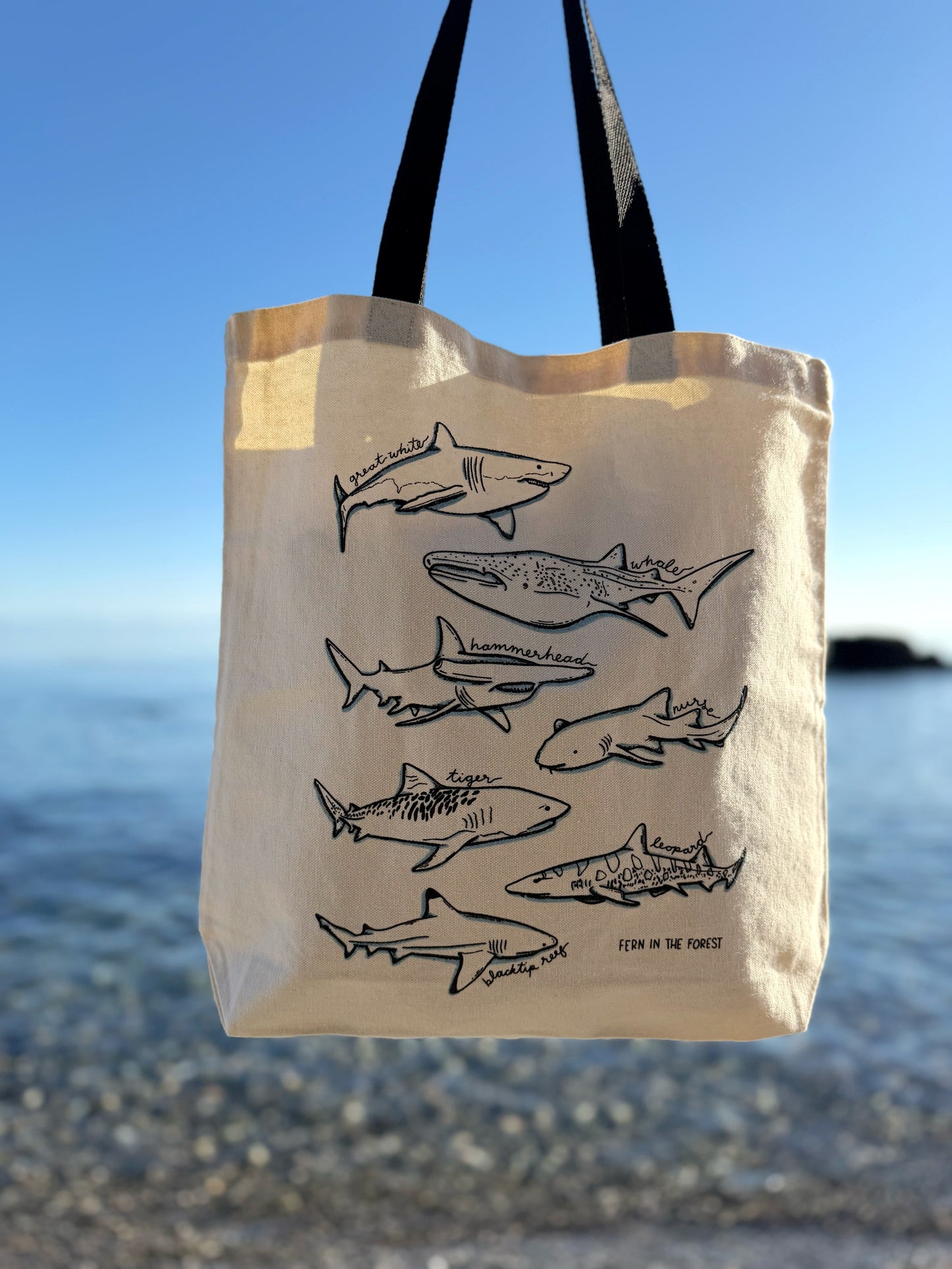 A canvas tote bag with black handles, featuring a black line art print of various sharks, hanging against a blurred beach and ocean background.