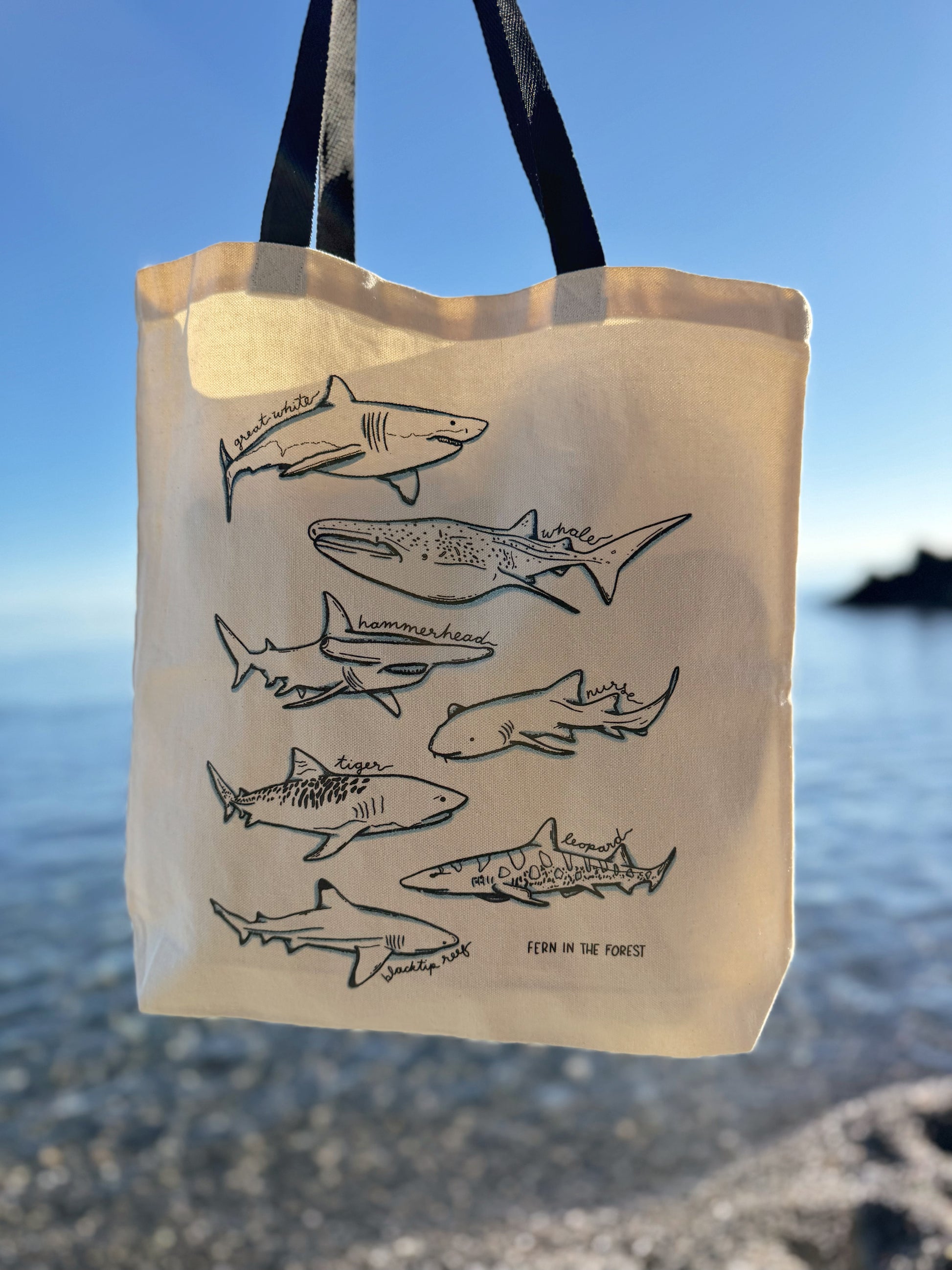 A cream-colored canvas tote bag with black handles, featuring a black line art print of various sharks. The bag is photographed outdoors against a blurred background of a blue sky and ocean water.