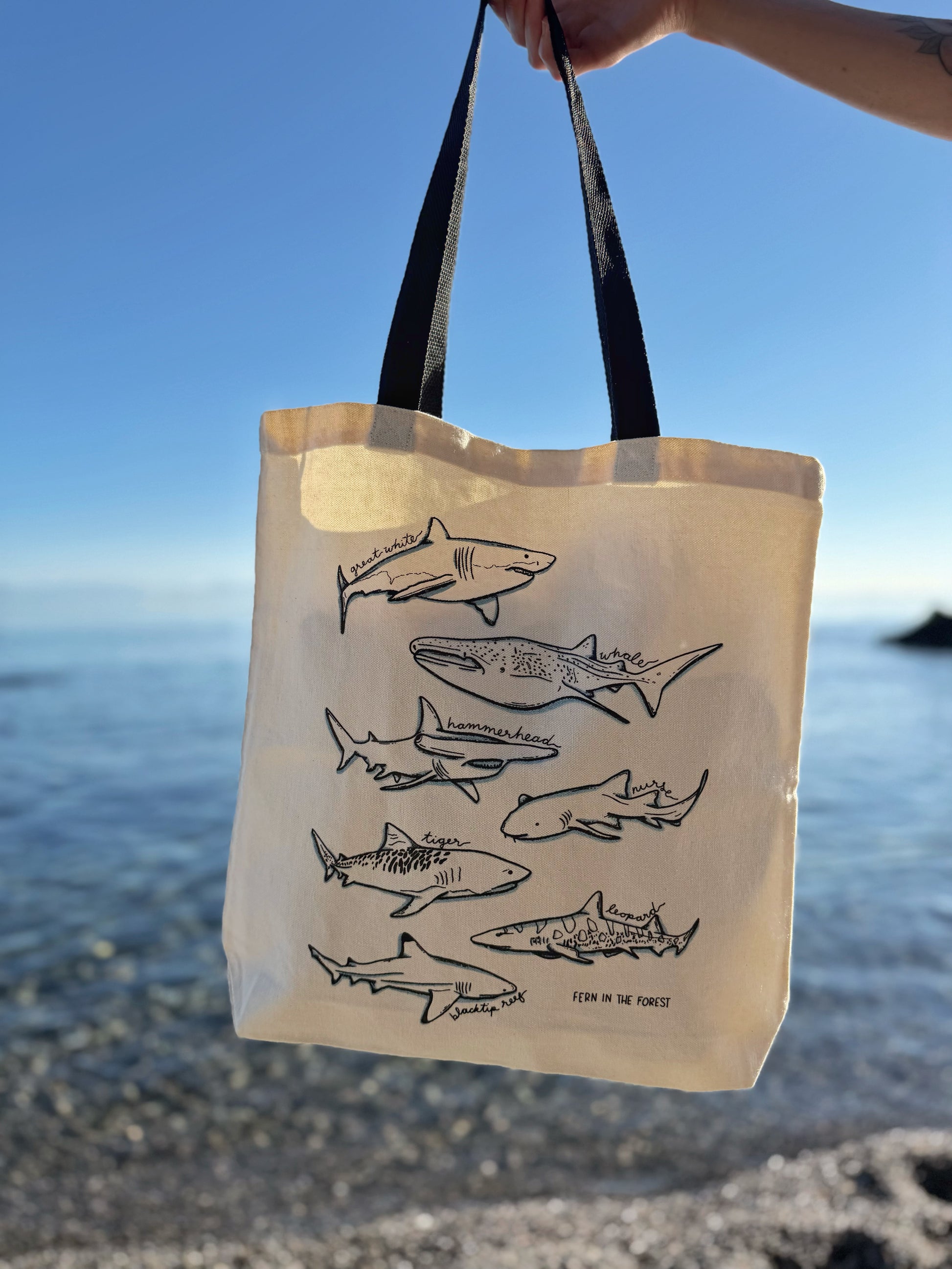 A cream-colored canvas tote bag with black handles, featuring a black line art print of various sharks with icy blue accents, held by a hand against a beach and ocean background.