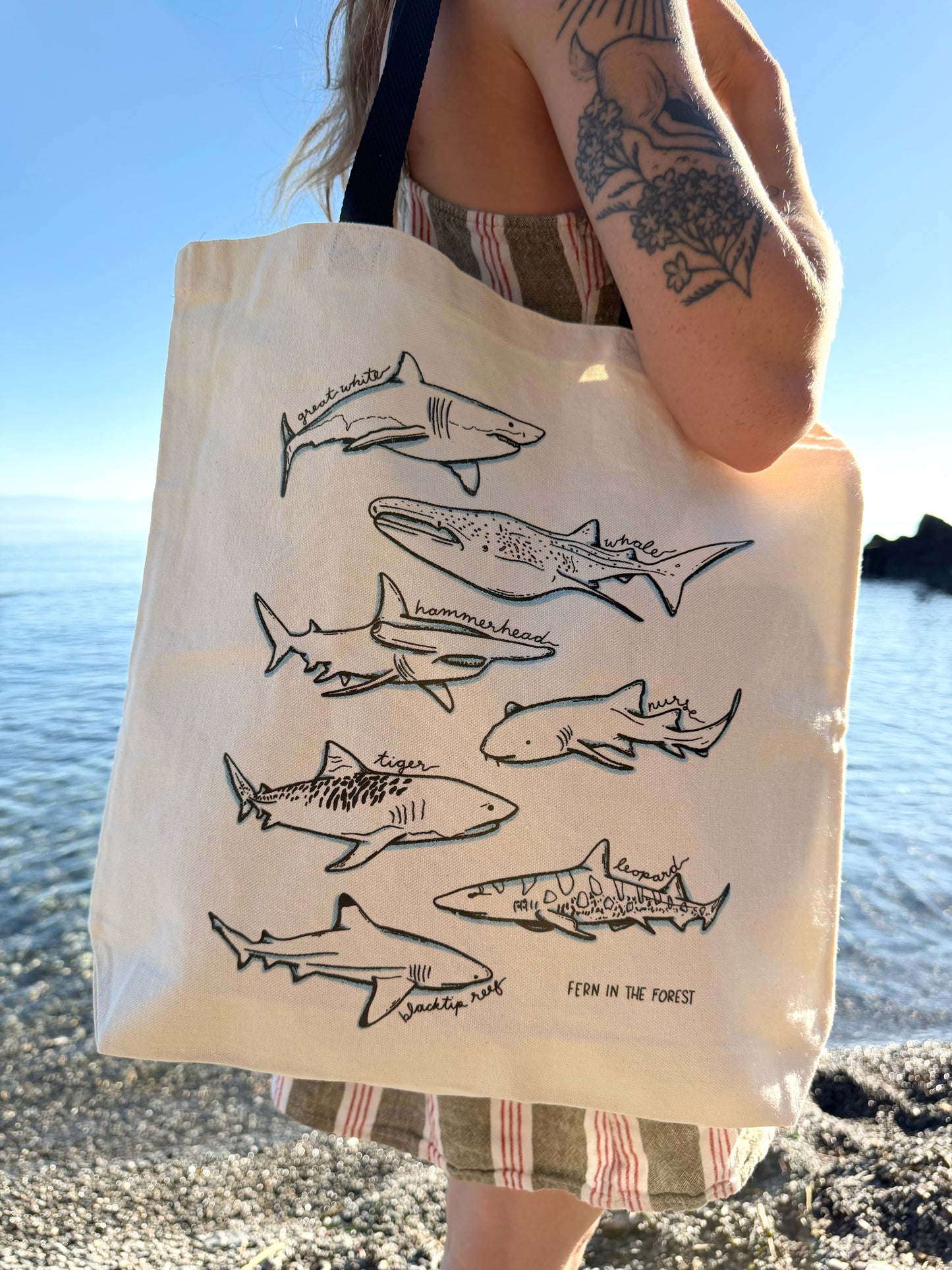 A person at the beach carrying a cream canvas tote bag printed with black line drawings of sharks with icy blue accents and the text 'FERN ON THE FOREST'.