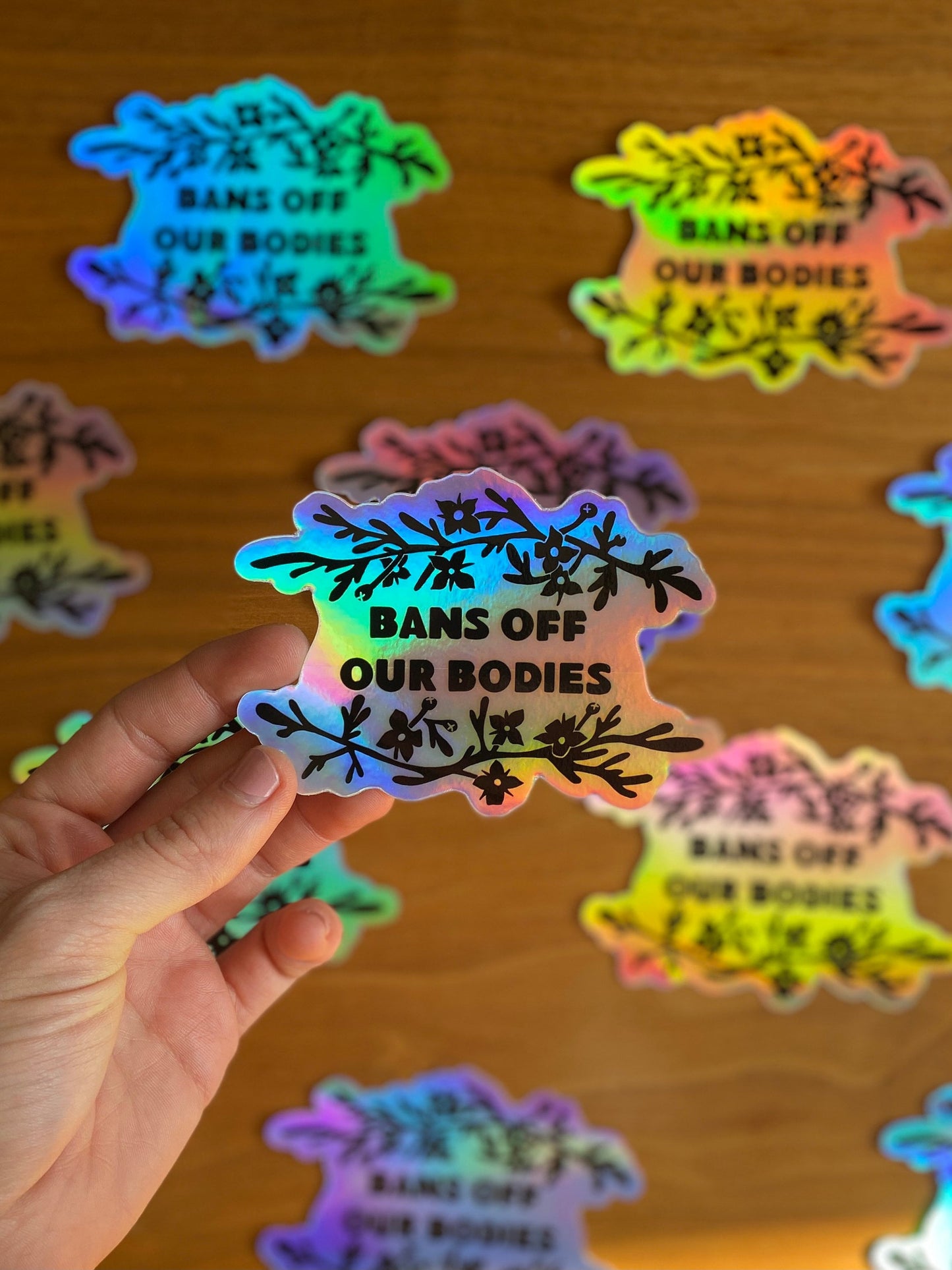 Holographic vinyl sticker that says "Bans off our bodies" with rue flower around it.