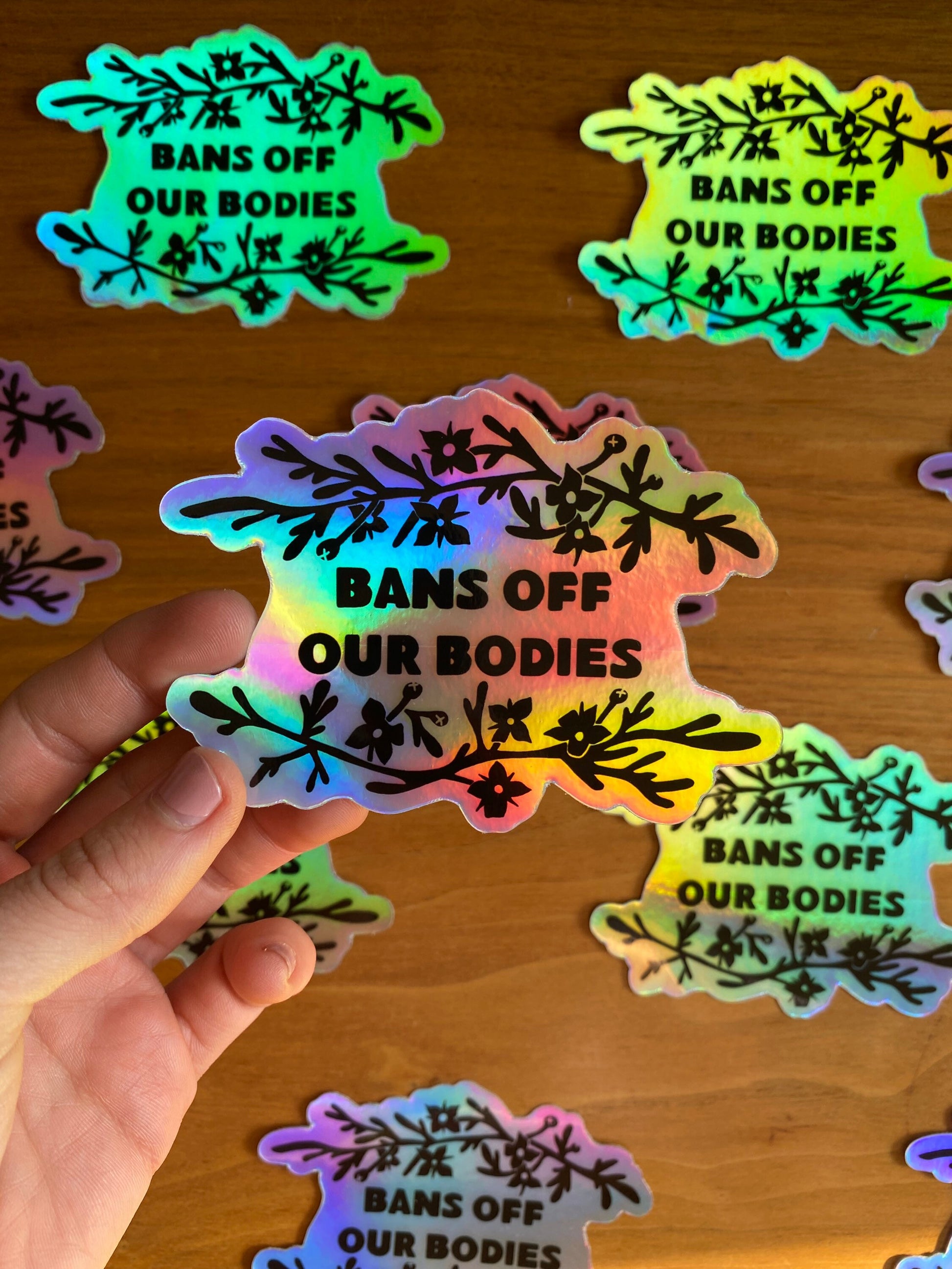 Holographic vinyl sticker that says "Bans off our bodies" with rue flower around it.